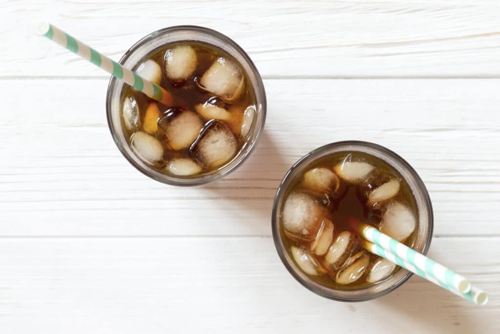 Black tea, coffee or cola with ice and paper straw in glass on white wooden background, top view. Two glass with drink on table. Summer beverage