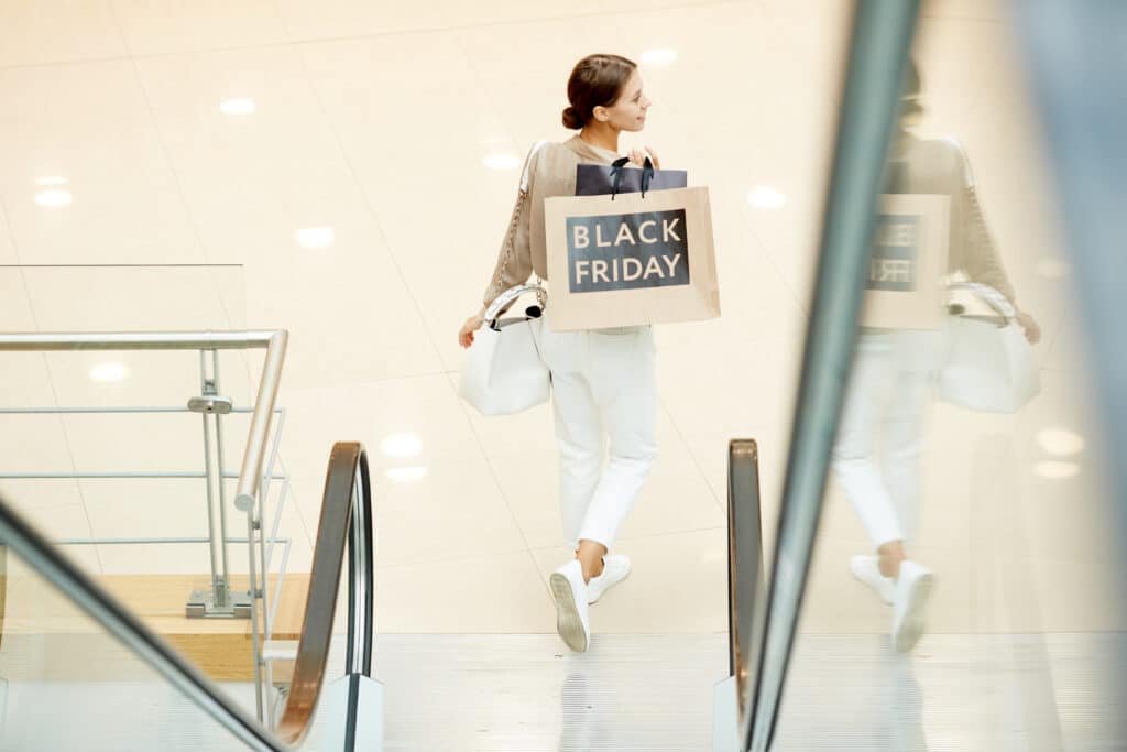 Vista trasera de una joven llevando bolsas de la compra mientras baja por las escaleras mecánicas de un centro comercial. Ofertas del Viernes Negro. 