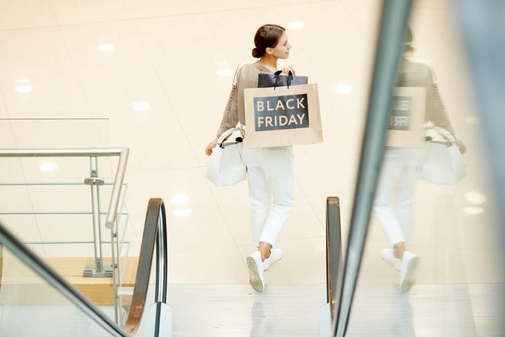 Rear view of young girl carrying shopping bags while moving down the escalator in the mall. Black Friday deals. 