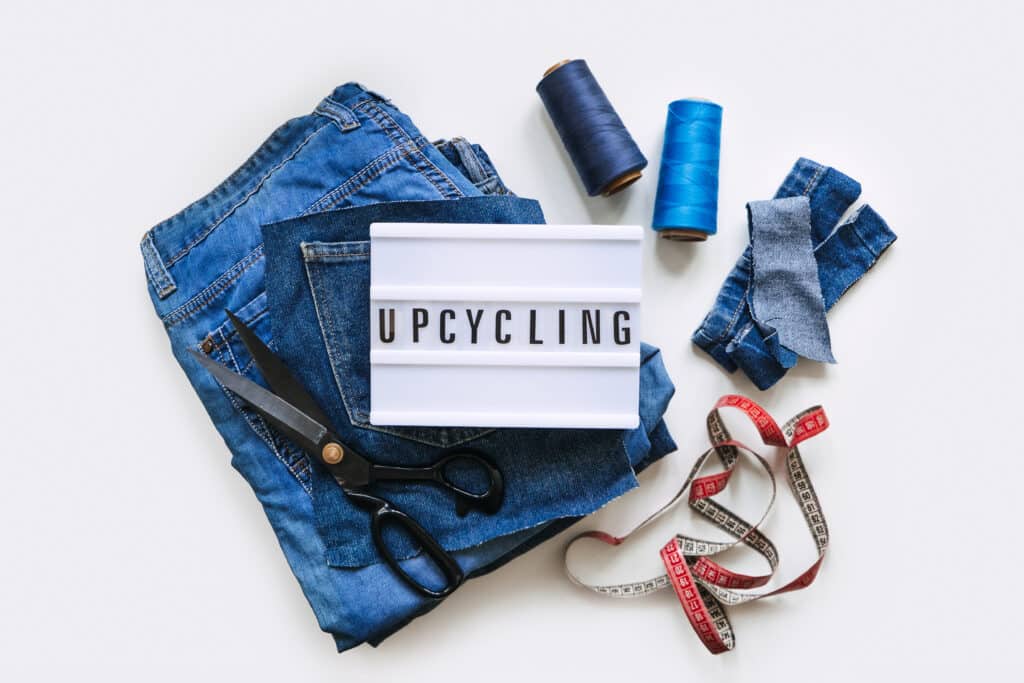 Denim Upcycling Ideas, Using Old Jeans, Repurposing Jeans, Reusing Old Jeans, Upcycle Stuff. Lightbox with text Upcycling, Stack of old blue jeans, scissors, thread and sewing tools in sewing studio.