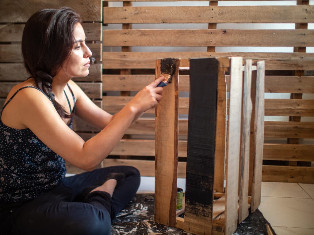 Young Hispanic Woman Dressed in Dark Clothes Paints a Wooden Box in Black, with a Pallet Background.

