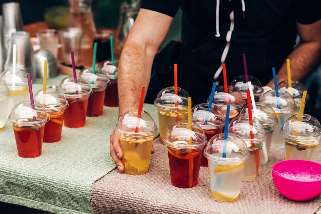hand serving lemonade with mint and lemons, space for text. street food festival. drink bar at reception, catering outdoors. summer picnic. colorful drinks in plastic cups with straws.