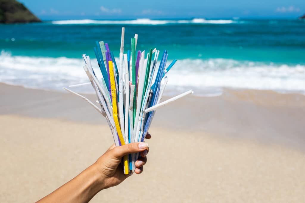 Hand holding heap of used plastic straws on background of clean beach and ocean waves. Plastic ocean pollution, environmental crisis. Say no plastic. Single-use plastic waste.
